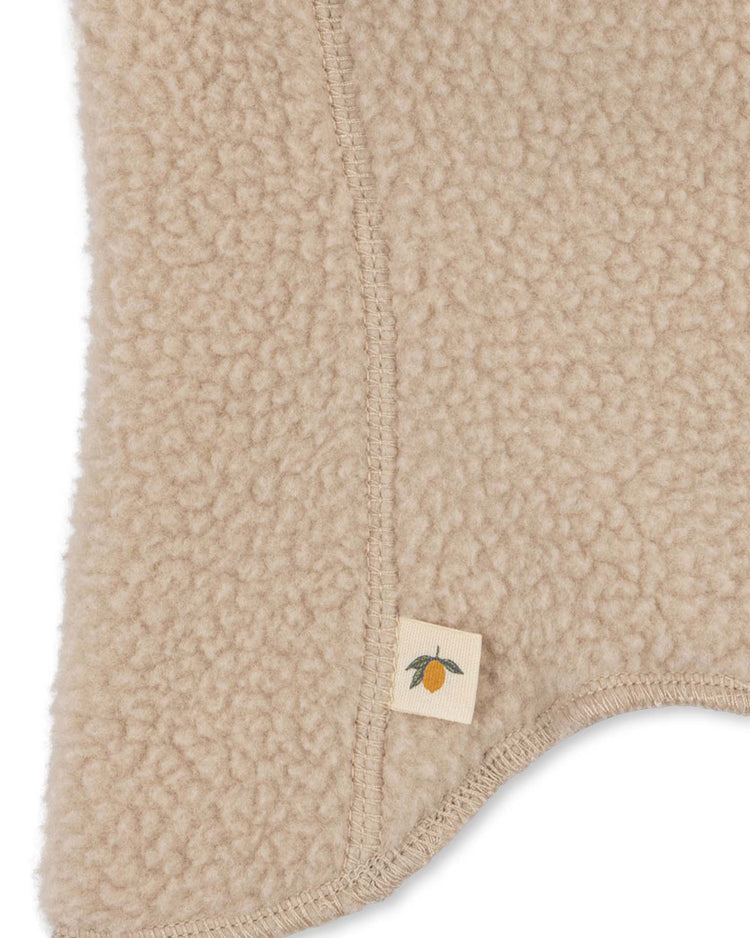 Close-up of beige, textured fleece fabric with visible stitching and a small fabric tag on the safa helmet in oxford tan by konges sløjd, featuring an illustration of an orange fruit with a green leaf.