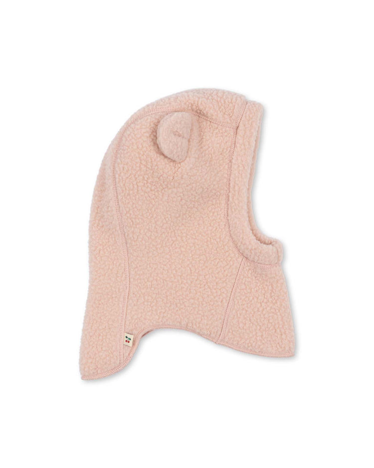 A light pink "safa helmet in rose smoke" by konges sløjd for children, featuring a bear ear detail on top and style code KS105768. The fleece hood covers the head, neck, and ears, with a small fabric tag near the bottom edge. Shown against a plain white background.