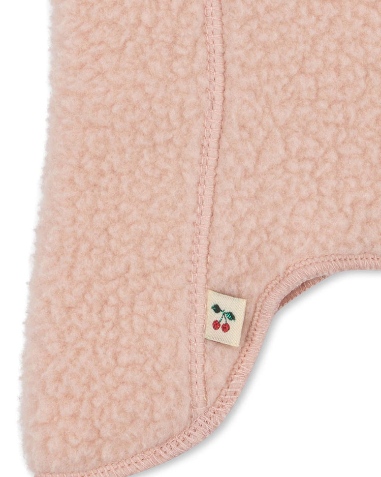 Close-up of the safa helmet in rose smoke by konges sløjd, featuring soft, pink, fleece-like fabric with visible stitching and a small white tag adorned with two red cherries and green leaves. The KS105768 design detail adds a unique touch to this cozy accessory.