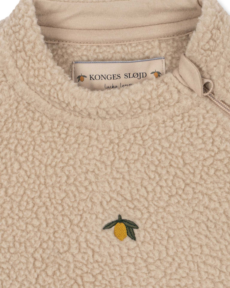 Close-up of the safa onesie in oxford tan by konges sløjd, a beige fleece-textured baby pullover featuring lemon embroidery on the chest and a branded label near the collar. Made from soft recycled polyester and OEKO-TEX certified materials, this cozy onesie also includes a visible zipper on the right side near the neckline.