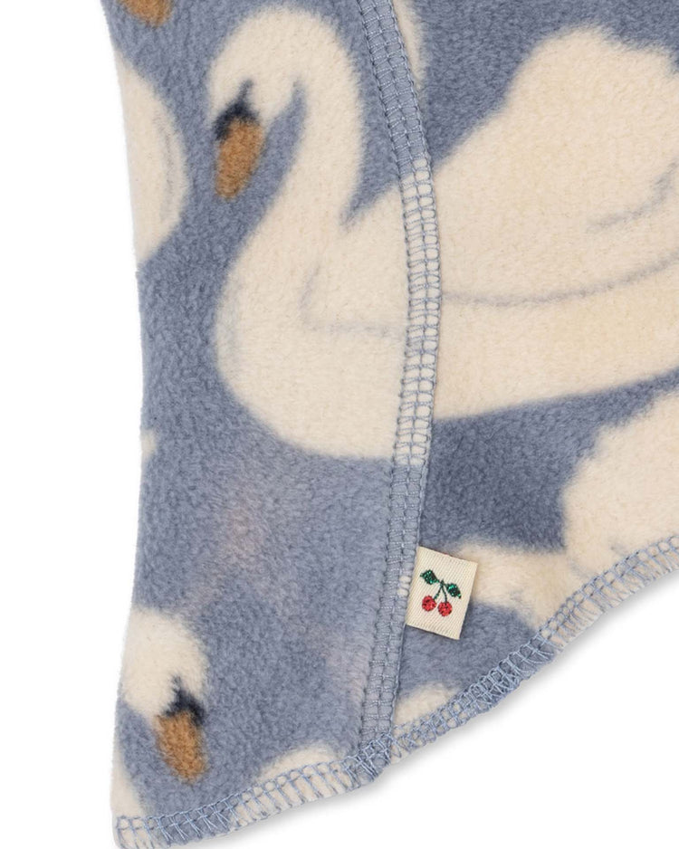 Close-up of the tavi fleece balaclava in swan by konges sløjd, showcasing soft fleece fabric with a white and grey-blue swan pattern, a small edge tag embroidered with red cherries, and the style code KS105772.