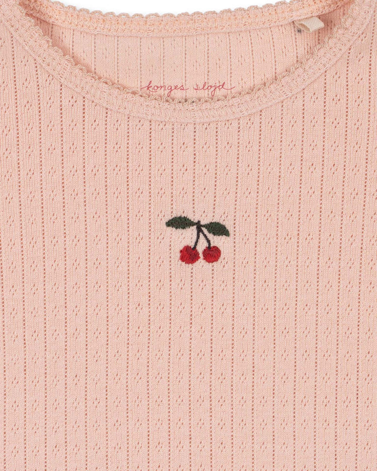 A close-up of the konges sløjd minnie blouse in pale blush shows its ribbed, light pink organic cotton fabric, highlighted by a delicate, embroidered cherry motif at the center and scalloped trim along the neckline—an ideal soft base layer, GOTS certified for sustainable comfort.