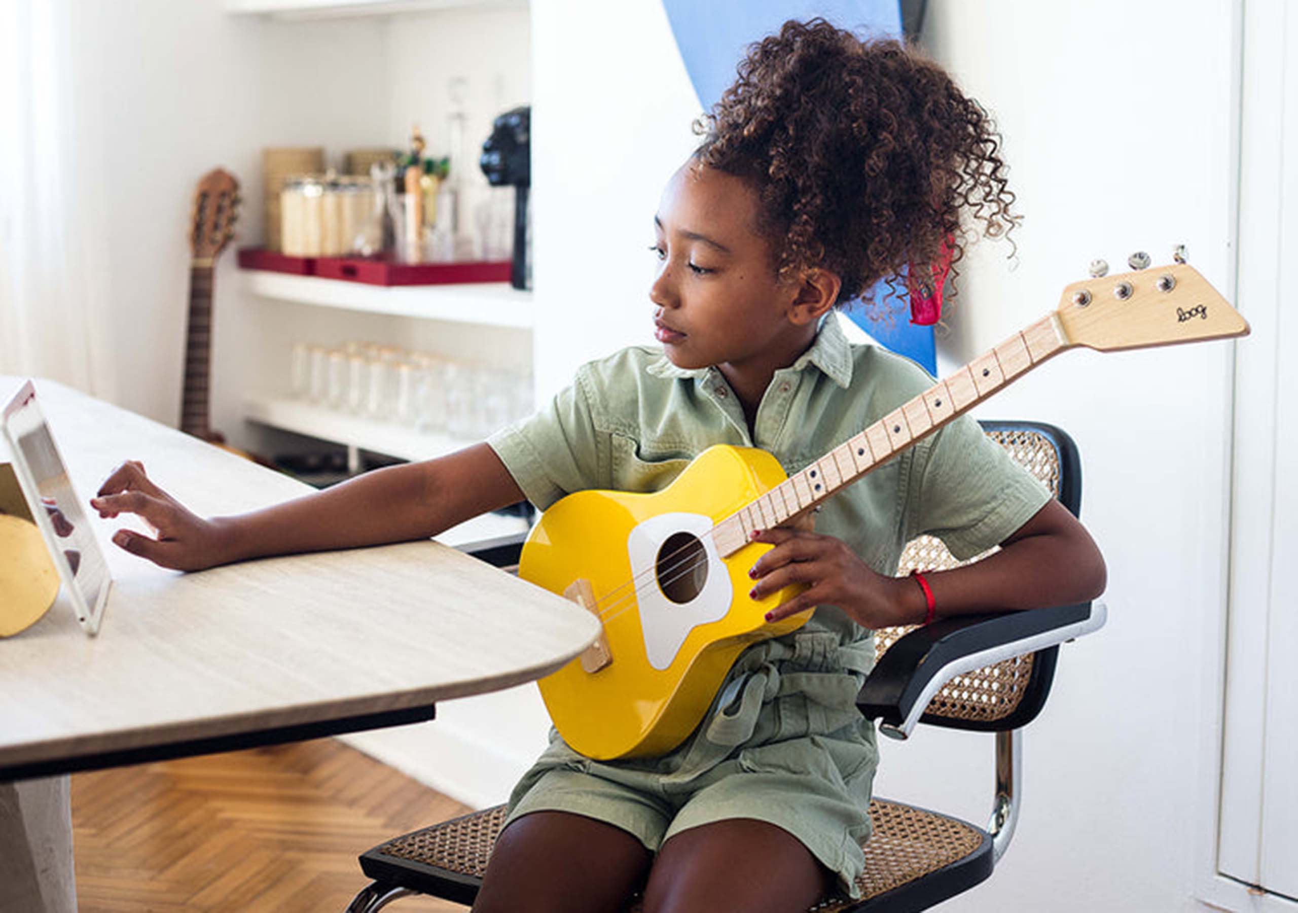A young child with a yellow guitar sits at a table, touching a tablet screen.