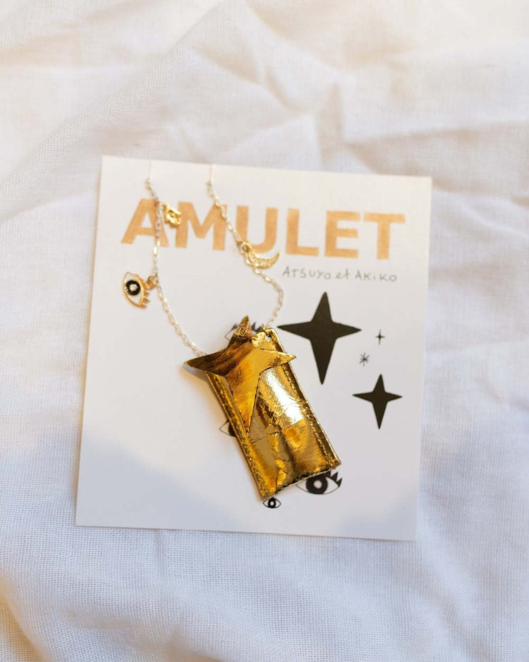 Gold-colored whistle pendant on a display card with the word "amulet" printed on it, now presented as an atsuyo et akiko amulet crystal necklace in foil gold.