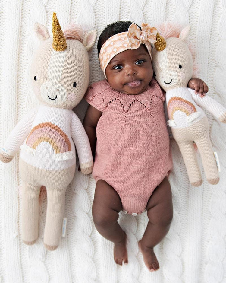 An infant wearing a headband lies next to two plush unicorn toys and a Cuddle + Kind Zara the Unicorn outfit.
