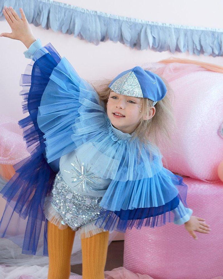 A little girl in a Meri Meri blue bird cape dress up set posing on a bed.