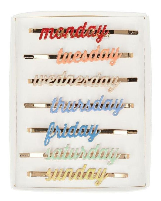 Seven Meri Meri enamel week day hair slides with the days of the week written on them, arranged in order on a white background.