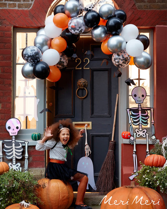 A child in a Halloween costume celebrates by a decorated doorway with meri meri halloween balloon garland kit, pumpkins, and skeleton decorations.