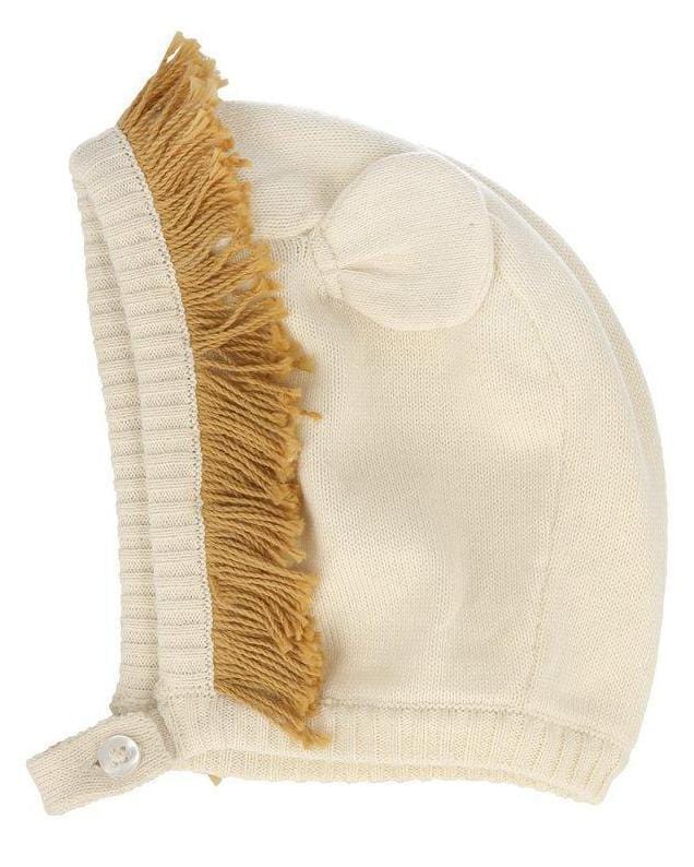 Cream-colored knitted lion baby bonnet with ear flaps and a fringed top, crafted from organic cotton by Meri Meri.