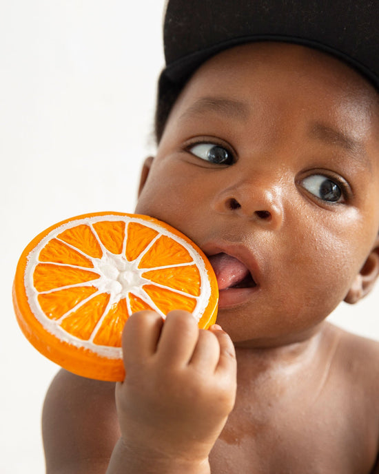 A baby wearing a black cap holds a clementino the orange from oli + carol, looking at it with curiosity, embodying a healthy lifestyle.