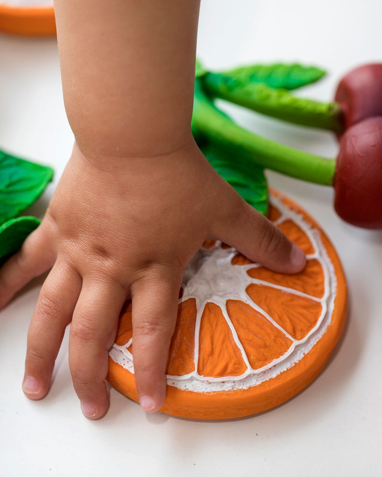 A child's hand pressing down on an oli + carol clementino the orange toy designed for teething.