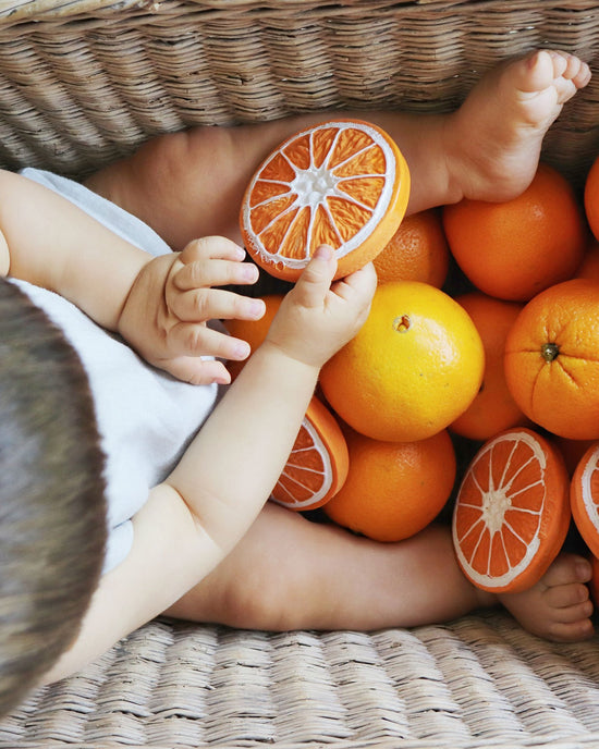 A baby teething with a basket of clementino the oranges, holding a half slice of clementino the orange from oli + carol.