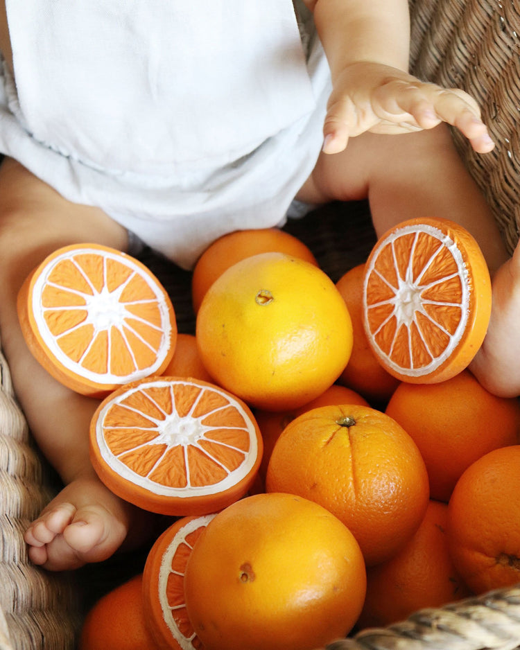 Toddler with a basket of fresh oranges, some cut in half to reveal the inside, playing with a oli + carol clementino the orange teething toy.