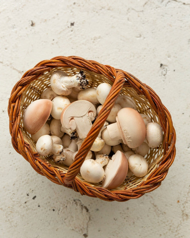 A wicker basket filled with oli + carol's manolo the mushroom sits beside a natural rubber baby toy on a textured surface.