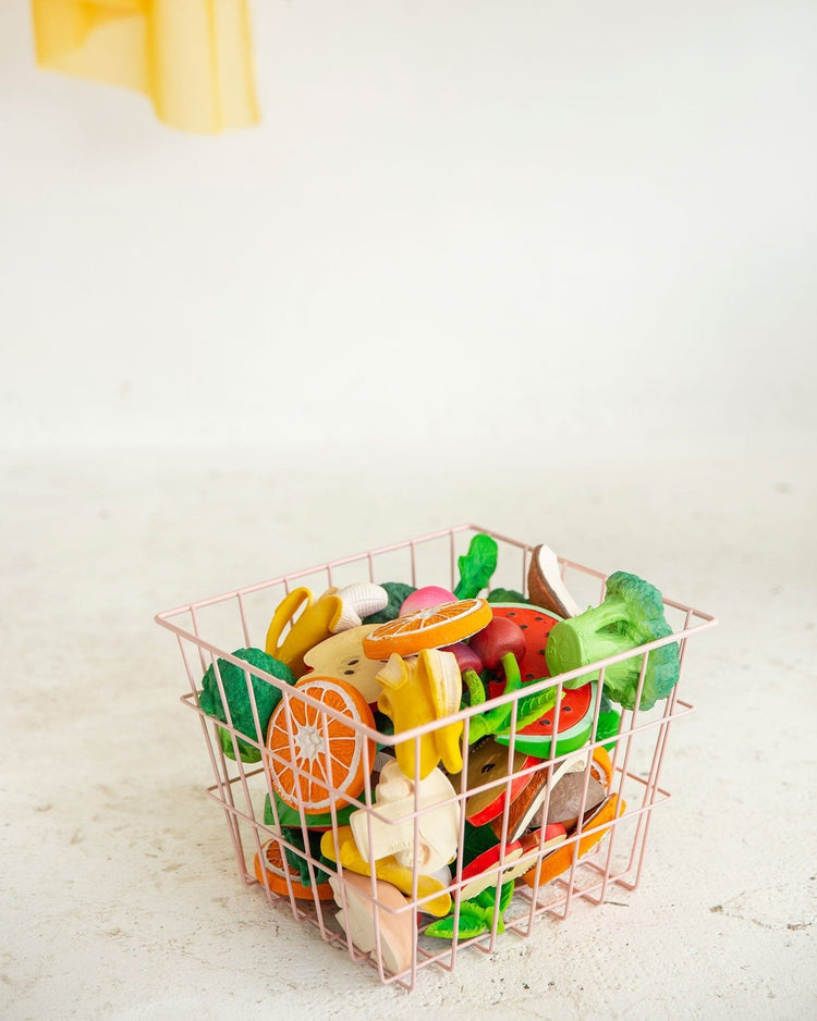 A shopping basket filled with assorted colorful oli + carol manolo the mushroom toy fruits and vegetables for baby teething, on a light background.