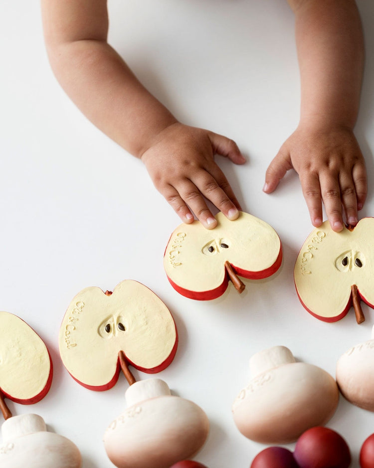 A baby's hands touching the Manolo the Mushroom model, designed as a natural rubber teething toy by oli + carol, amidst whole apples and apple halves on a white surface.