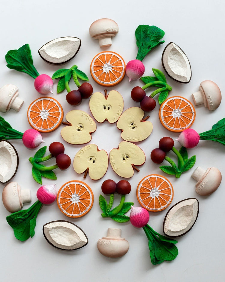 A symmetrical arrangement of assorted fruits and vegetables on a white background, including a oli + carol manolo the mushroom natural rubber teething baby toy.