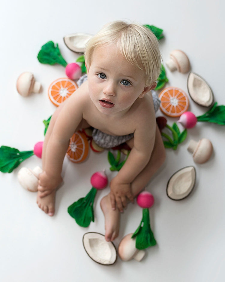 Toddler surrounded by colorful teething toy manolo the mushroom fruits and vegetables from oli + carol on a white background.