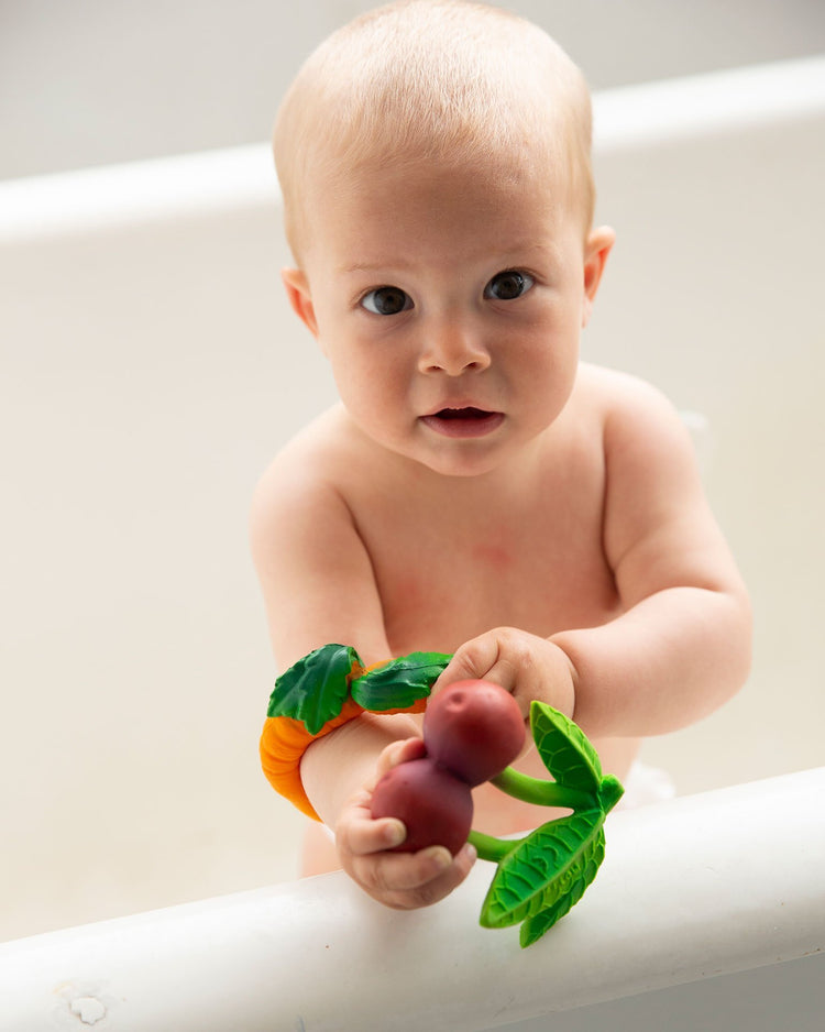 Infant holding a mery the cherry teething baby toy fruit while standing behind a railing from oli + carol.