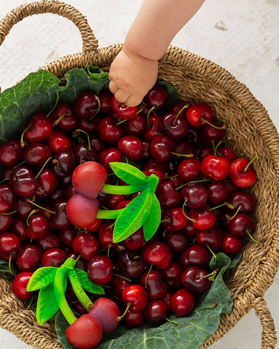 A child's hand reaching into a wicker basket full of oli + carol's mery the cherry eco-toy cherries with green leaves.