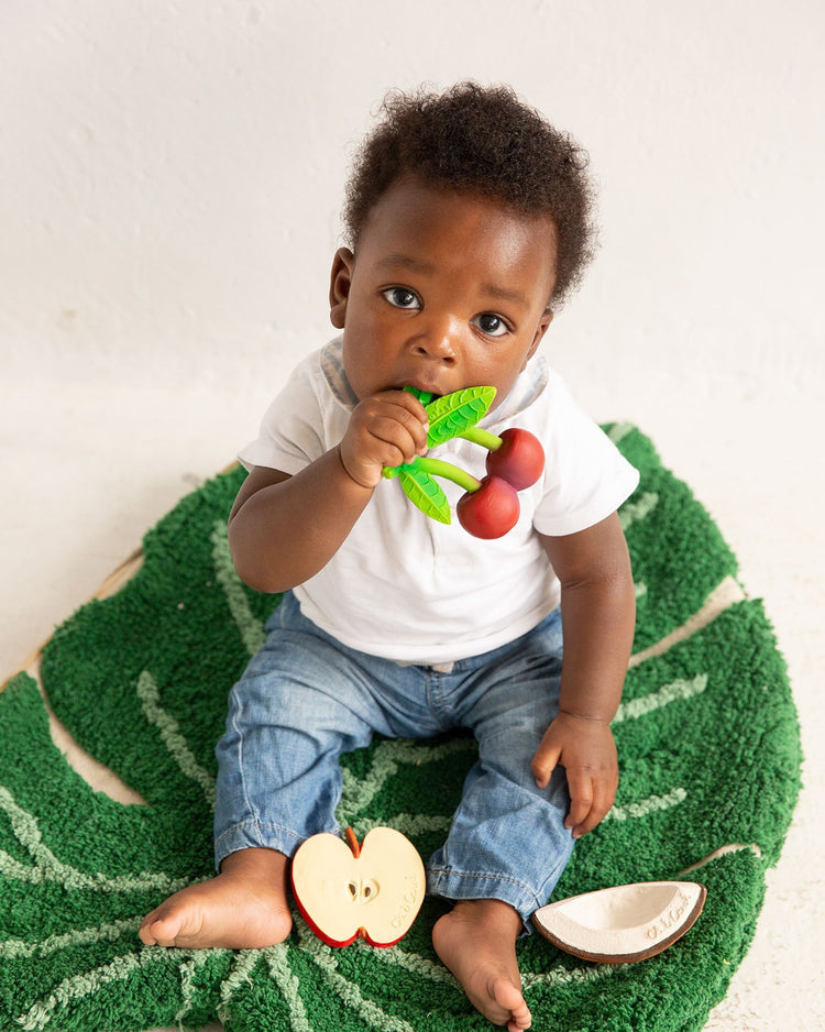 Toddler sitting on a green playmat, chewing on an oli + carol mery the cherry baby toy fruit while surrounded by more toy fruits.