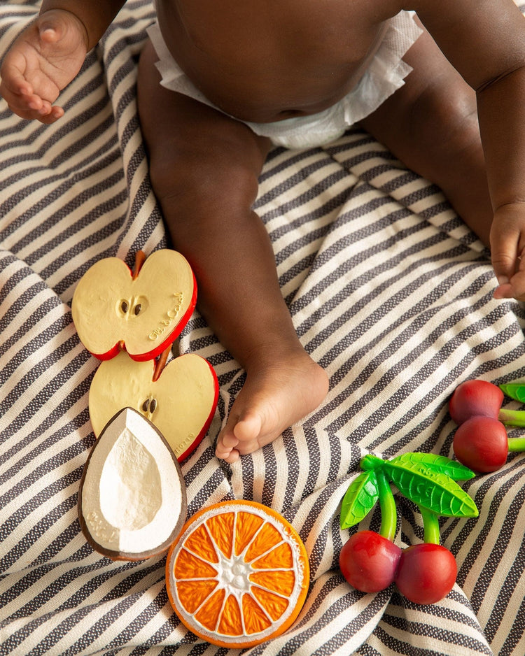 An overhead view of a baby in a diaper seated on a striped blanket, surrounded by oli + carol's mery the cherry wooden fruit teething toys.