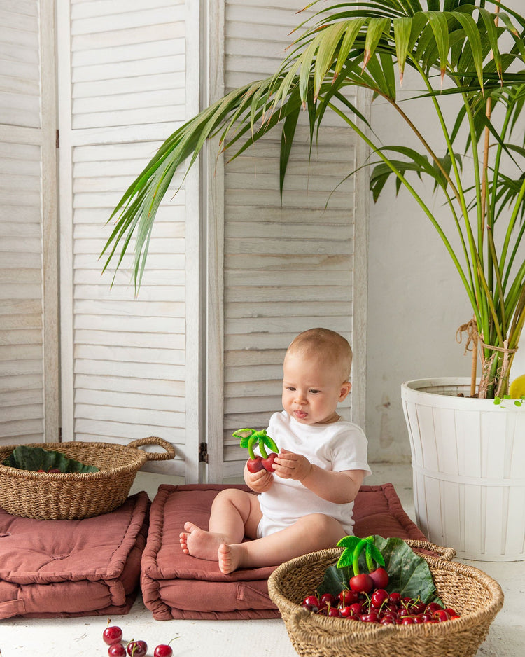 Toddler sitting on a mat indoors, playing with an oli + carol mery the cherry baby toy next to a basket of cherries.
