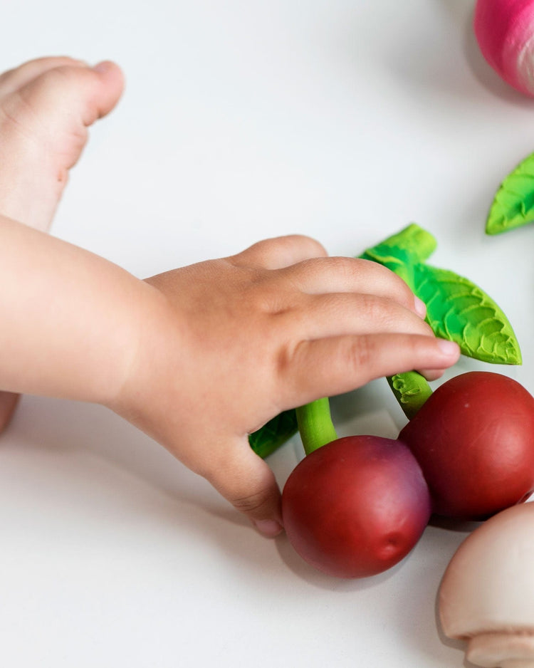 A toddler's hand reaching for the oli + carol mery the cherry on a white surface.