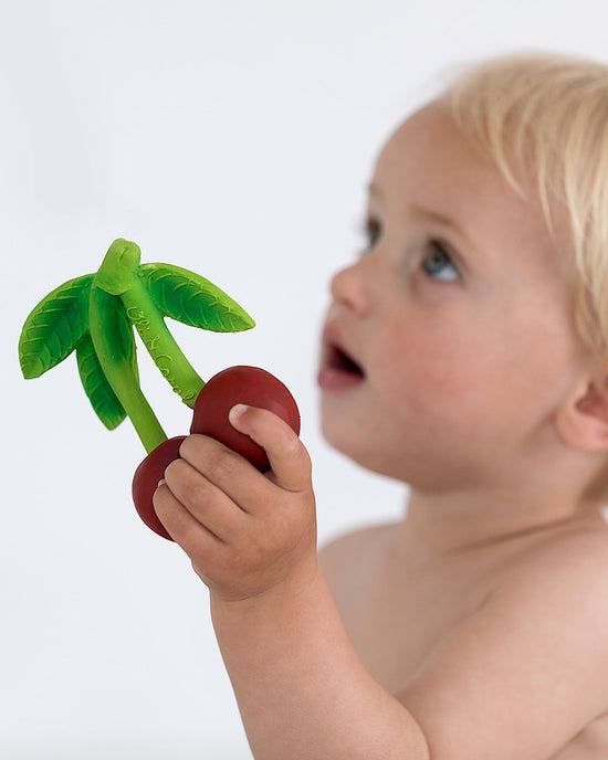 Toddler looking at an oli + carol mery the cherry toy apple with surprise and curiosity.