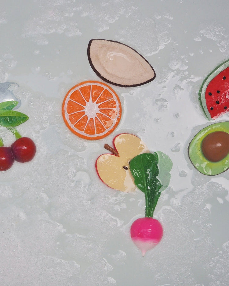 Flat lay of colorful mery the cherry baby toy teething soaps by oli + carol on a sudsy surface.
